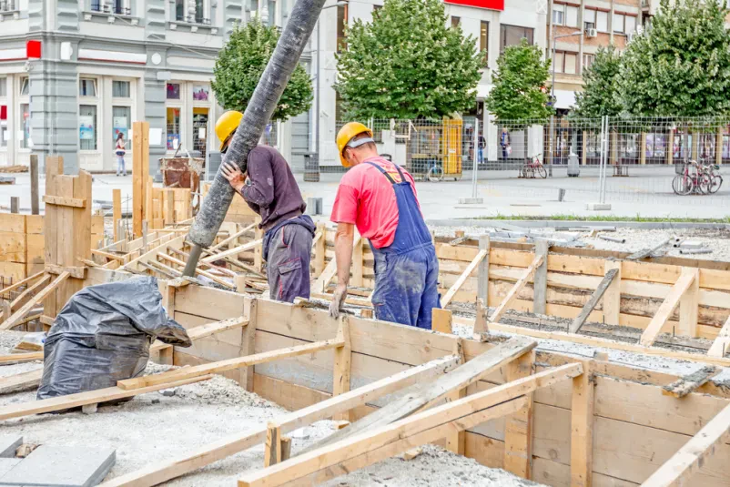 Gradbeni delavci s pomočjo cevi usmerjajo svež beton v leseni opaž, skrbno pripravljen za vlivanje. Prikaz delo z lesom pri betoniranju.