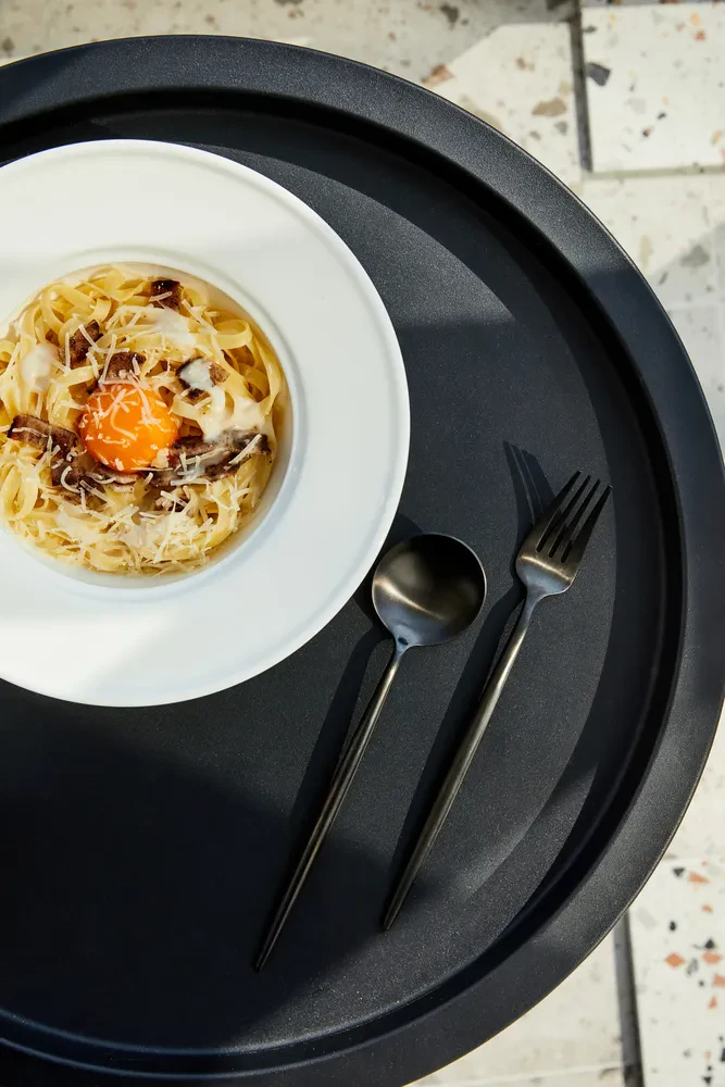 Razkošna porcija testenin carbonara s hrustljavo slanino, rumenjakom in parmezanom, servirana na temni mizi.
