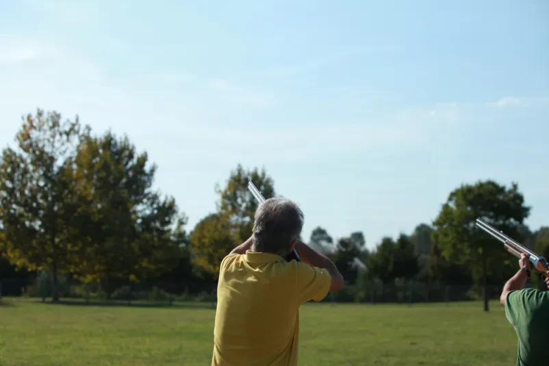 Skupina strelcev na glinaste golobe Skupina strelcev z vso opremo, vključno z zaščitnimi očali, merijo s puškami proti nebu in čakajo na izstrelitev glinastega goloba med tekmovanjem.
