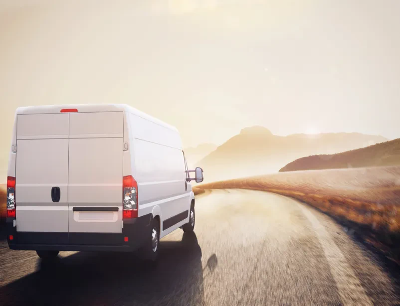 Velik beli tovornjak se pelje po cesti v naravni pokrajini. Predstavlja medkrajevni transport in prevoz tovora.