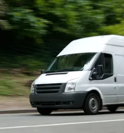 Beli dostavni kombi se hitro premika po cesti, z zabrisanim ozadjem zelenja. Poudarja hitrost in učinkovitost kombi prevozov.