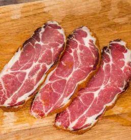 Od blizu posnete, tanko narezane rezine sušene svinjske vratovine, skrbno položene na leseno rezalno desko.