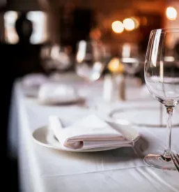 Elegantno pogrnjena restavracijska miza z belimi prtički, sijočim priborom in praznim kozarcem za vodo.