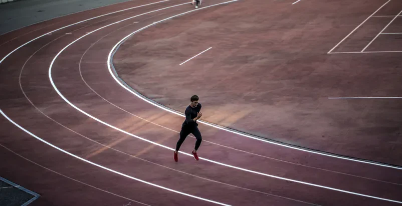 Atlet teče po stezi, posneto od zgoraj Prizor od zgoraj prikazuje atleta med šprintom na rdeči atletski stezi. Poudarjena sta hitrost in dinamika gibanja med treningom.