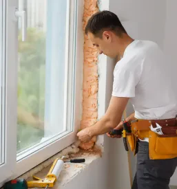 Moški industrijski delavec v delovni obleki in rokavicah skrbno vgrajuje okno na gradbišču, kar prikazuje profesionalno in ugodno montažo oken v Sloveniji.
