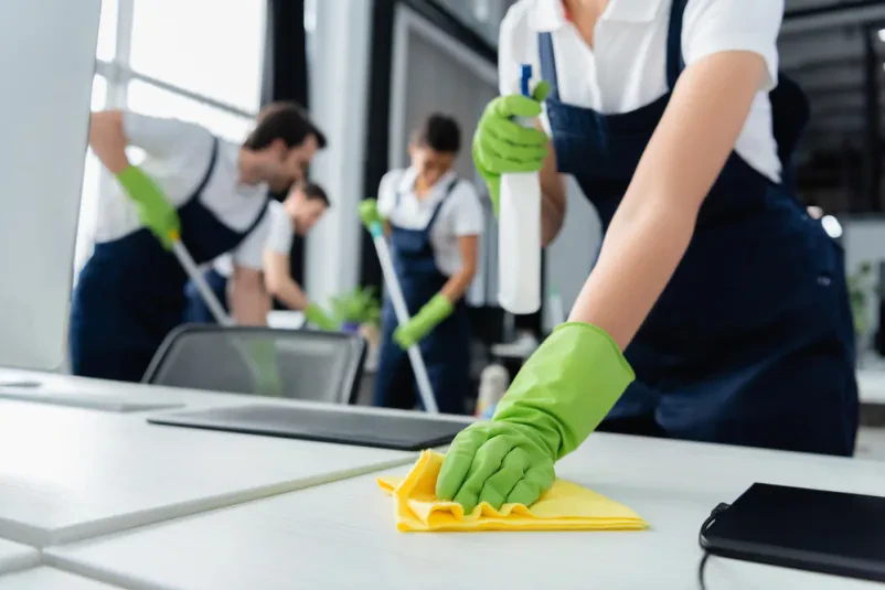 Čistilka z rokavico v roki drži stekleničko detergenta in krpo, pripravljena za temeljito čiščenje mize v sodobni pisarni.