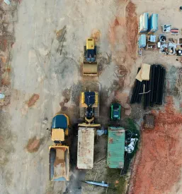 Zračni posnetek gradbišča, ki prikazuje buldožer, tovornjak kiper, kompaktor za zemljo in drenažne cevi za pripravo terena.
