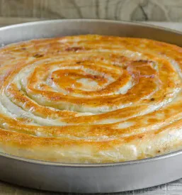 Okrogel burek, lepo pečen do zlato rjave barve, postrežen v kovinskem pekaču na leseni podlagi. Svež in topel.