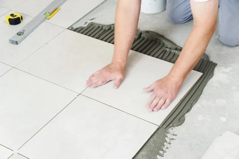 Delavčeve roke pritiskajo keramično ploščico na pripravljena tla, s čimer zagotavljajo pravilno namestitev in ravno površino.