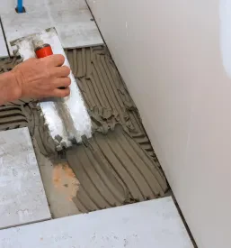 Roke delavca z gladilko nanašajo lepilo na tla kopalnice, ključen korak pri skrbni pripravi za polaganje keramičnih ploščic.