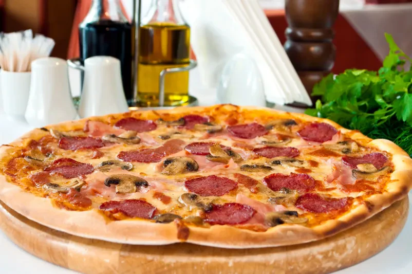 Izjemno okusna, sveže pečena pica s tankimi rezinami salame, gobami in svežo baziliko, ki čaka na postrežbo. Hrustljava skorja in bogata obloga sta znak kakovostne kuhinje.