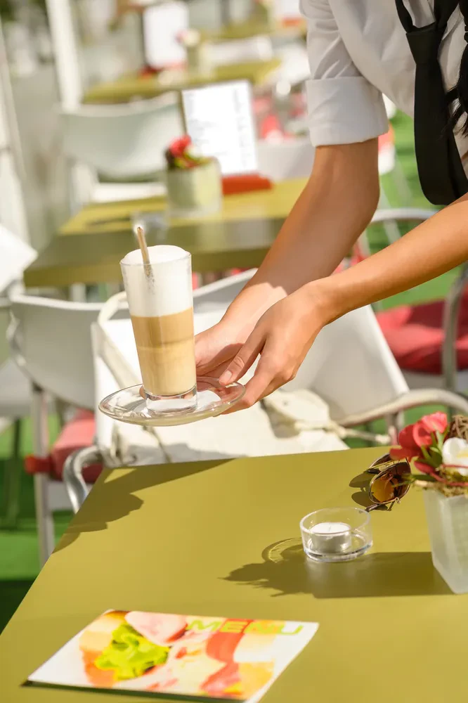 Natakarica postreže latte Natakarica s skrbnostjo postreže sveže pripravljeno kavo latte na leseni mizi zunanji terasi, poudarjajoč kakovost pijač.