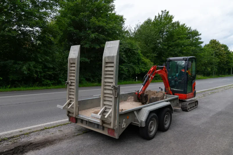 Rdeč bager je naložen na prikolico ob cesti, pripravljen za prevoz. Rampi sta spuščeni, žlica bagra pa se dotika tal.