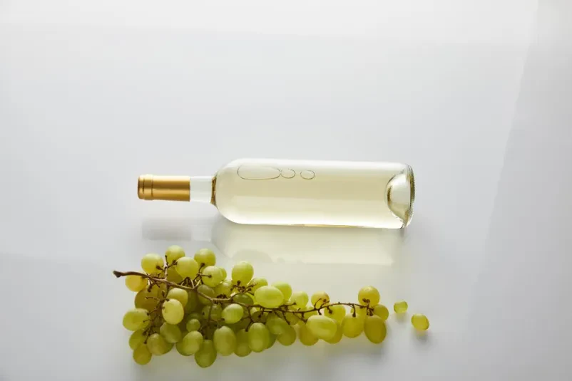 Pogled od zgoraj na steklenico kakovostnega belega vina, postavljeno ob svežem grozdju na čistem belem ozadju. Poudarja svežino in kakovost pijače.
