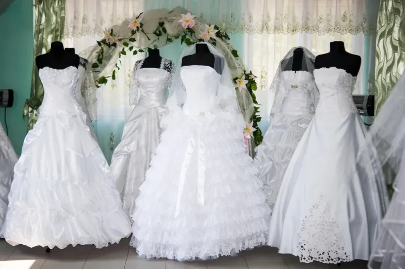 Elegantne poročne obleke, obešene v svetlem poročnem salonu, čakajo na neveste za Prodajo poročnih in svečanih oblek Pomurje.