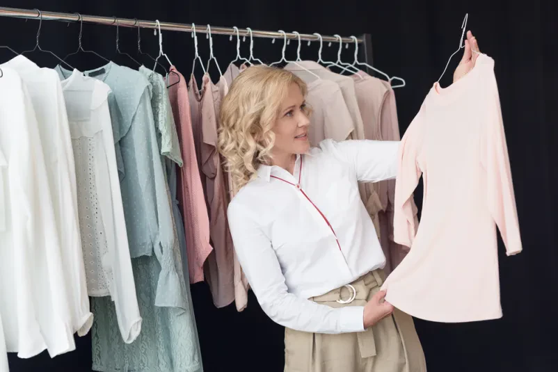 Ženska skrbno izbira oblačila v modni trgovini Ženska skrbno pregleduje in izbira oblačila na obešalnikih. Išče nove modne kose med pestrim asortimanom ženskih oblačil.
