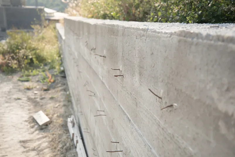 Bližnji posnetek del v teku, kjer iz sveže ulitega betonskega podpornega zidu štrlijo armaturne palice. To kaže na fazo gradbenih izkopov in zidarskih del pred dokončanjem objekta.