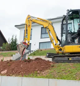 Majhen rumen gosenični bager ureja travnato pobočje nad temelji novega betonskega podpornega zidu med prenovo. Stroji so ključni za učinkovito izvedbo zemeljskih del in pripravo terena.
